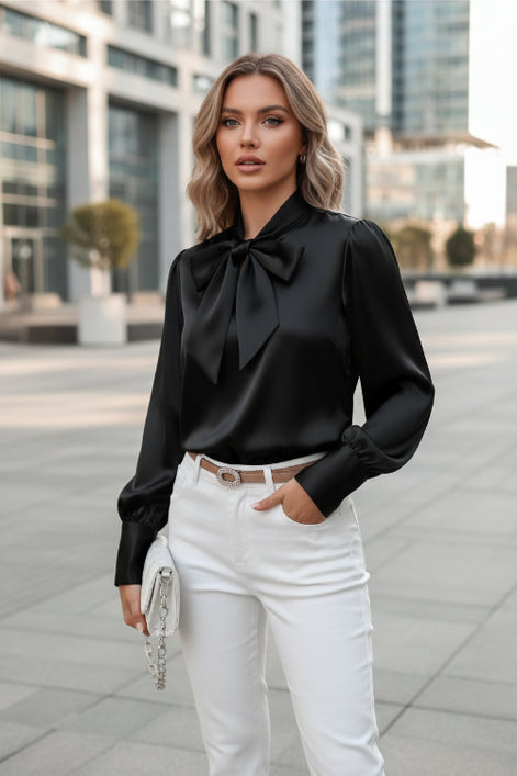 Black satin blouse with a large bow tie worn by a person, sitting on a white chair.