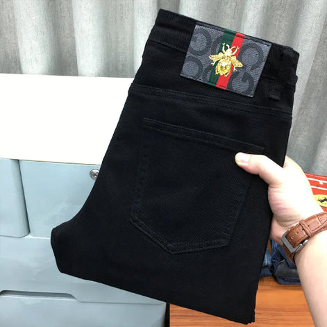 Black Gucci jeans folded and held by a hand, showing the iconic rear pocket and the signature Gucci web stripe with a bee patch at the waistband.