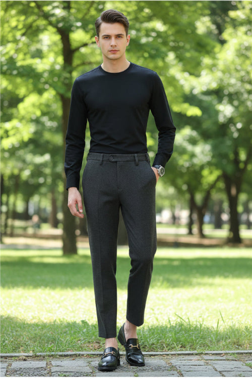 A person wearing dark gray textured slim-fit trousers, paired with a black long-sleeve top and black loafers, standing in front of a modern glass-paneled wooden wall.