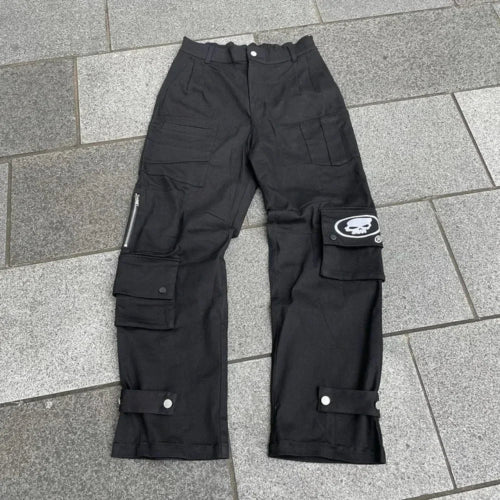Front view of black cargo pants laid flat on gray tiles, featuring multiple flap pockets, a zippered pocket, a white skull graphic, and snap-button straps near the hems.⠋