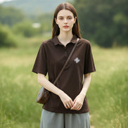 A woman wearing a dark brown short-sleeve polo shirt with a small white geometric logo on the left chest, paired with light gray bottoms and carrying a brown shoulder bag.