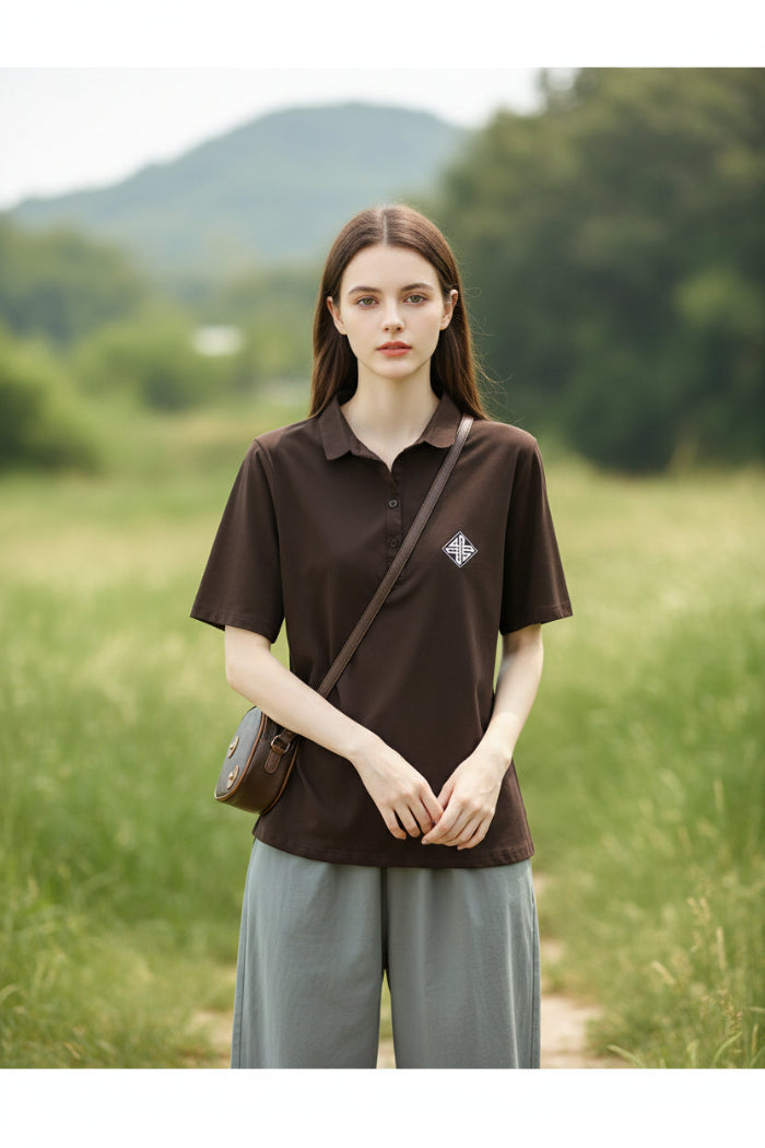 A woman wearing a dark brown short-sleeve polo shirt with a small white geometric logo on the left chest, paired with light gray bottoms and carrying a brown shoulder bag.