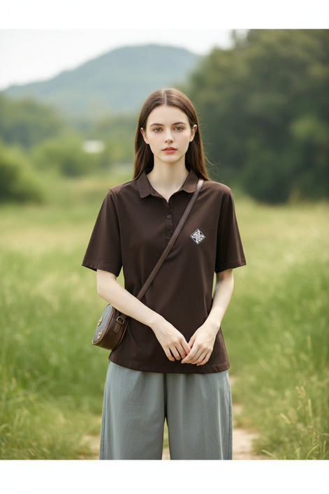 A woman wearing a dark brown short-sleeve polo shirt with a small white geometric logo on the left chest, paired with light gray bottoms and carrying a brown shoulder bag.