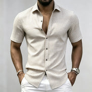 A man wearing a beige and white checkered short-sleeve button-up shirt with white pants, shown from the chin down against a light gray background.