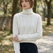 A person wearing a fuzzy off-white turtleneck sweater with long sleeves, paired with a long silver beaded necklace and dark pants, standing indoors.