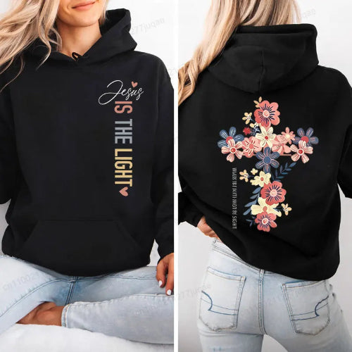 A woman modeling a black pullover hoodie shown from both front and back. The front has white and gold text reading 'Jesus IS THE LIGHT' with small pink heart accents.