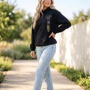 A woman modeling a black pullover hoodie shown from both front and back. The front has white and gold text reading 'Jesus IS THE LIGHT' with small pink heart accents.