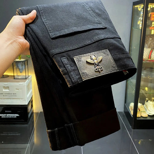A hand holds up a pair of black denim jeans, showing the back pocket with a large decorative metal winged skull patch. 