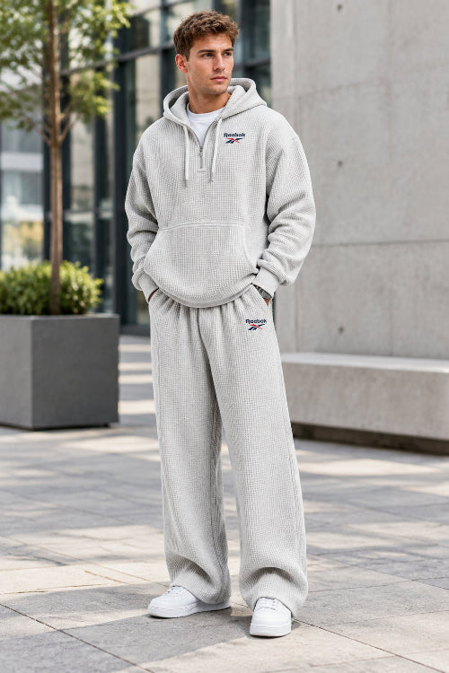 A man wearing a light gray waffle-knit Reebok tracksuit, featuring a pullover hoodie and matching sweatpants, against a plain white background.