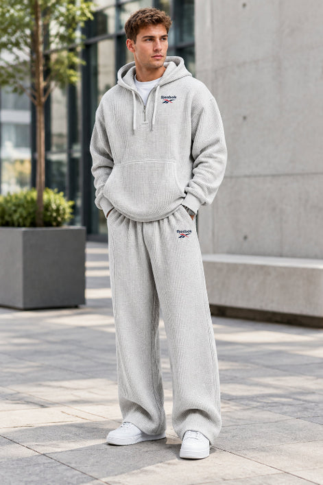 A man wearing a light gray waffle-knit Reebok tracksuit, featuring a pullover hoodie and matching sweatpants, against a plain white background.
