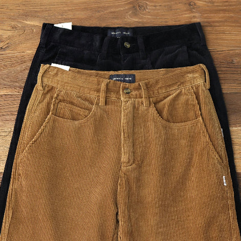 Three pairs of corduroy pants in brown, black, and tan are stacked flat on a wooden surface. The front pair is tan, the middle pair is black, and the back pair is a warm brown.