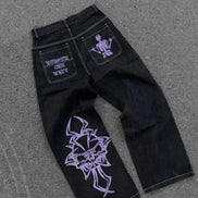 Back view of black wide-leg jeans laid flat on a gray surface, featuring purple lettering and a skeleton graphic on the back pockets and a large purple cross-style design on one leg.