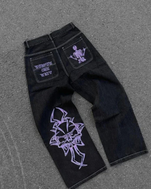 Back view of black wide-leg jeans laid flat on a gray surface, featuring purple lettering and a skeleton graphic on the back pockets and a large purple cross-style design on one leg.