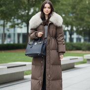 A woman takes a mirror selfie in a clothing store while wearing a long brown puffer coat 