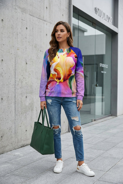 A woman wearing a vibrant, multicolored long-sleeve sweatshirt with a large floral print, paired with blue jeans, holding a bouquet of white roses.