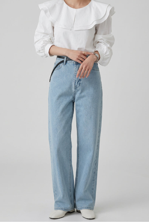 A woman wearing a white long-sleeve blouse with a large ruffled collar and light blue high-waisted jeans, standing indoors.
