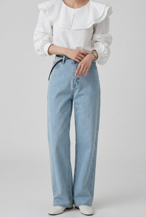 A woman wearing a white long-sleeve blouse with a large ruffled collar and light blue high-waisted jeans, standing indoors.