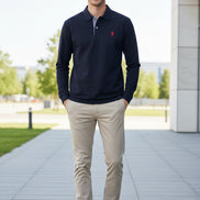 Navy blue polo shirt with red logo held by a hand, with more shirts in the background.