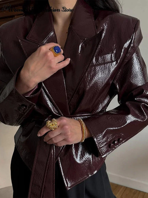 Close-up of a glossy dark brown belted jacket showing wide lapels, a chest welt pocket, textured fabric and buttoned cuffs