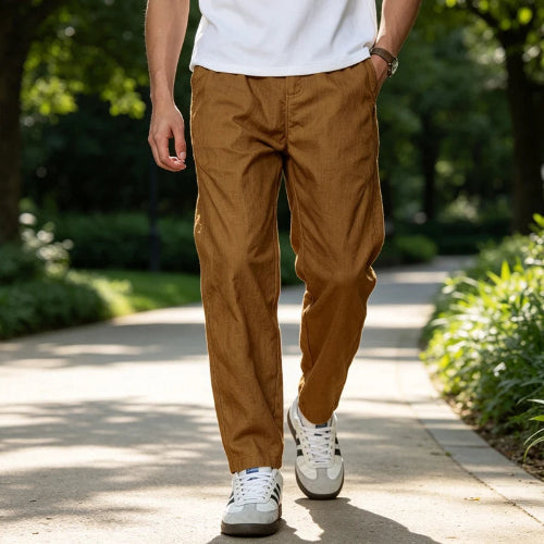 A person walking on a sunlit park path wearing slim brown pants, a white T-shirt, and white sneakers with black stripes.