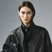 Close-up front view of a woman wearing a black faux leather jacket with a zipped inner collar and layered outer collar against a gray background