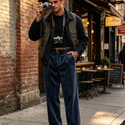A man stands on a city sidewalk wearing a black long-sleeve shirt, olive green utility vest, dark blue corduroy pants, and black-and-white loafers, 