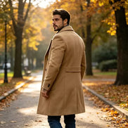 A man wearing a mid-length camel brown coat and blue jeans stands on a tree-lined path with fallen leaves, looking back over his shoulder.