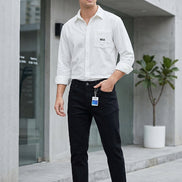 Black slim-fit jeans hanging on a metal rack, displayed alongside a white button-up shirt in a styled retail setting with a gray wall, floor lamp, plant, and storage boxes.