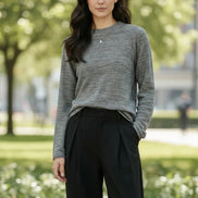 A woman wearing a gray long-sleeve knit sweater and black pants, standing indoors near a window with soft light. The sweater appears to be the product being shown.