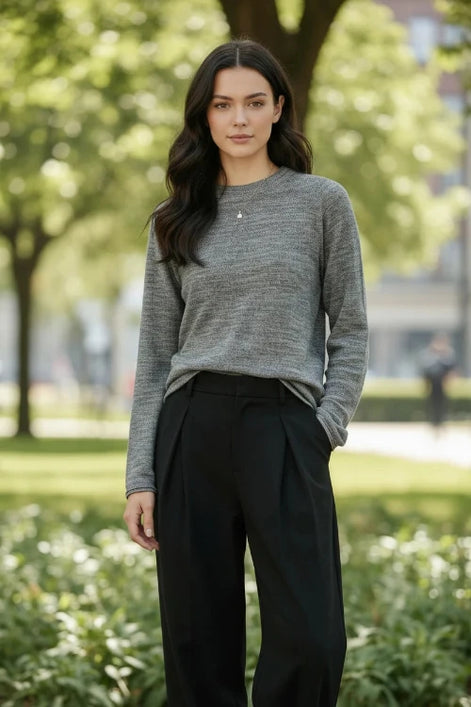 A woman wearing a gray long-sleeve knit sweater and black pants, standing indoors near a window with soft light. The sweater appears to be the product being shown.