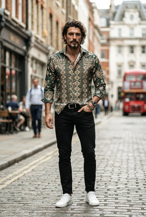 A man wearing a long-sleeve button-up shirt with an intricate dark green, beige, and brown paisley and geometric pattern, paired with black trousers and a black belt.