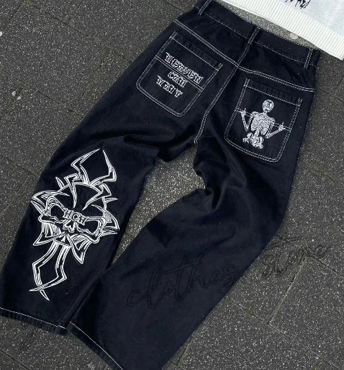 Back view of black wide-leg jeans laid flat on a gray surface, featuring red lettering and a skeleton graphic on the back pockets and a large red cross-style design on one leg.