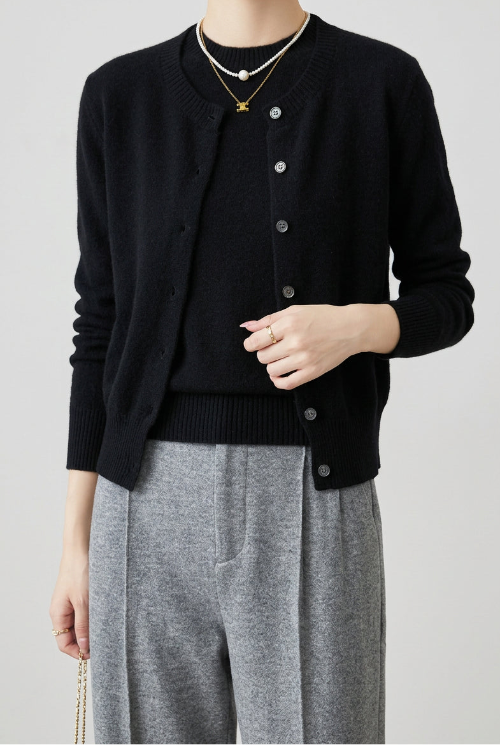 A person wearing a black knit cardigan with silver buttons over a black top and gray pants, standing indoors near a brown leather chair. 