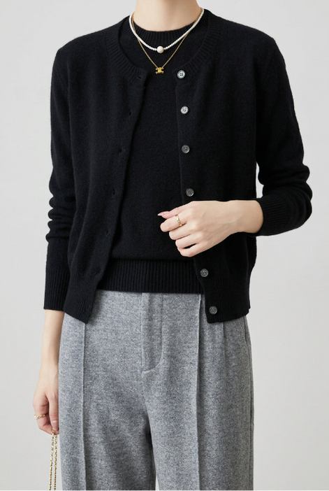 A person wearing a black knit cardigan with silver buttons over a black top and gray pants, standing indoors near a brown leather chair. 