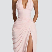 A model wearing a light pink halter-neck maxi dress with a deep plunging neckline, ruched bodice, draped skirt details, 