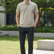 A close-up of a beige short-sleeve polo shirt on a black hanger, held by a hand to show the fabric texture. The shirt features a subtle landscape pattern on the front and a small circular logo on the chest pocket.