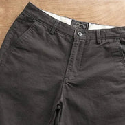 A pair of dark charcoal gray chino pants laid flat on a light brown wooden surface, showing the waistband, belt loops, button, and front pockets.