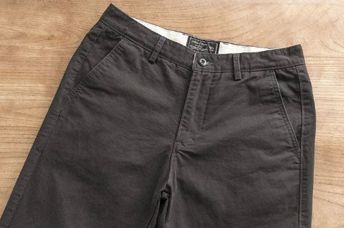 A pair of dark charcoal gray chino pants laid flat on a light brown wooden surface, showing the waistband, belt loops, button, and front pockets.