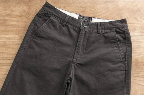 A pair of dark charcoal gray chino pants laid flat on a light brown wooden surface, showing the waistband, belt loops, button, and front pockets.
