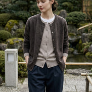 A person wearing a dark brown button-front cardigan over a light beige cardigan and white shirt, paired with dark pants, in an indoor setting with a warm lamp on a table.