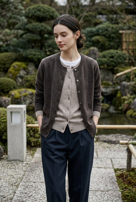 A person wearing a dark brown button-front cardigan over a light beige cardigan and white shirt, paired with dark pants, in an indoor setting with a warm lamp on a table.