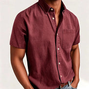 A man wearing a short-sleeve button-up shirt in a dusty burgundy or maroon color, paired with blue jeans. 