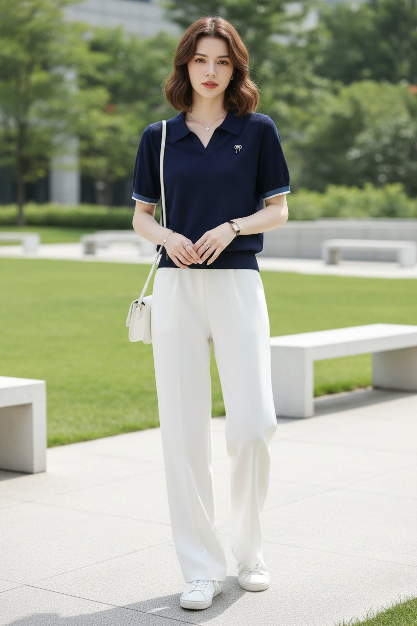 A woman wearing a navy blue short-sleeve polo shirt with a textured knit fabric, a small bow pin on the chest, and striped trim on the collar and sleeve cuffs, paired with white bottoms.