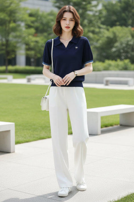 A woman wearing a navy blue short-sleeve polo shirt with a textured knit fabric, a small bow pin on the chest, and striped trim on the collar and sleeve cuffs, paired with white bottoms.