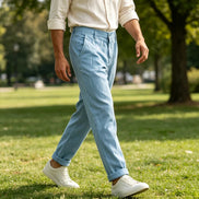 A man walking outdoors in a park wearing light blue chino pants, a cream linen shirt, and white sneakers.