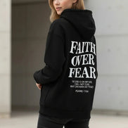 Black hoodie with 'Faith Over Fear' text on a wooden surface with shoes and headphones.