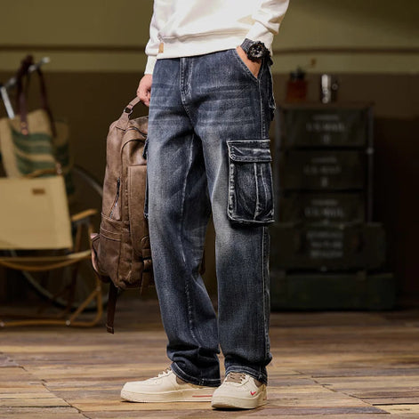 Front-angle view of a model wearing faded dark blue denim cargo pants with a large flap thigh pocket, holding a brown backpack and wearing light-colored sneakers
