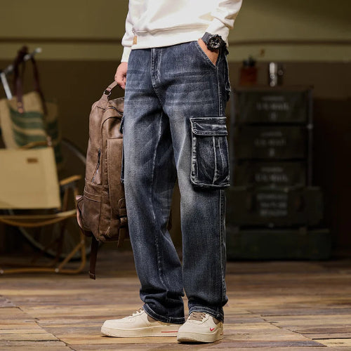 Front-angle view of a model wearing faded dark blue denim cargo pants with a large flap thigh pocket, holding a brown backpack and wearing light-colored sneakers