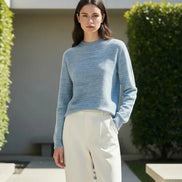 A woman wearing a light blue heathered knit mock-neck sweater and white pants, shown from the lips down in a softly lit indoor setting.