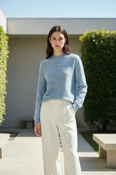 A woman wearing a light blue heathered knit mock-neck sweater and white pants, shown from the lips down in a softly lit indoor setting.